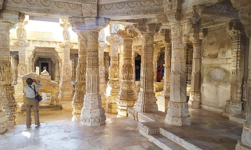 A person photographing the intricate carvings inside a sunlit, ancient stone temple with numerous pillars and detailed architectural elements, capturing one of Rajasthan's architectural wonders.