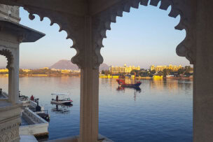 View across a lake framed by an ornate architectural arch, revealing the wonders of serene boats on the water and splendid buildings along the shoreline in the distance with a majestic mountain backdrop.