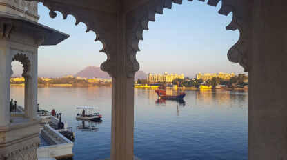 View across a lake framed by an ornate architectural arch, revealing the wonders of serene boats on the water and splendid buildings along the shoreline in the distance with a majestic mountain backdrop.