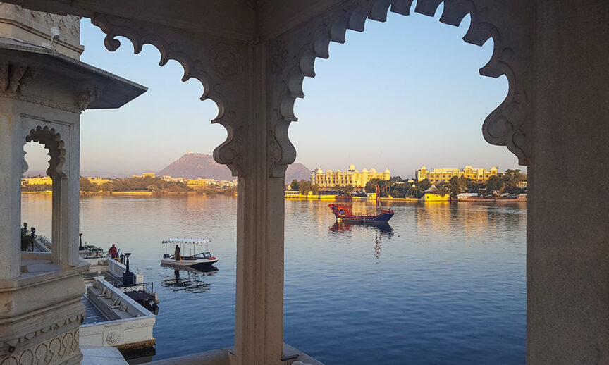 View across a lake framed by an ornate architectural arch, revealing the wonders of serene boats on the water and splendid buildings along the shoreline in the distance with a majestic mountain backdrop.