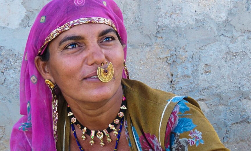 A woman in traditional attire, wearing a pink headscarf, ornate nosering, and colorful necklace, stands against a textured wall, embodying the wonders of Rajasthan.