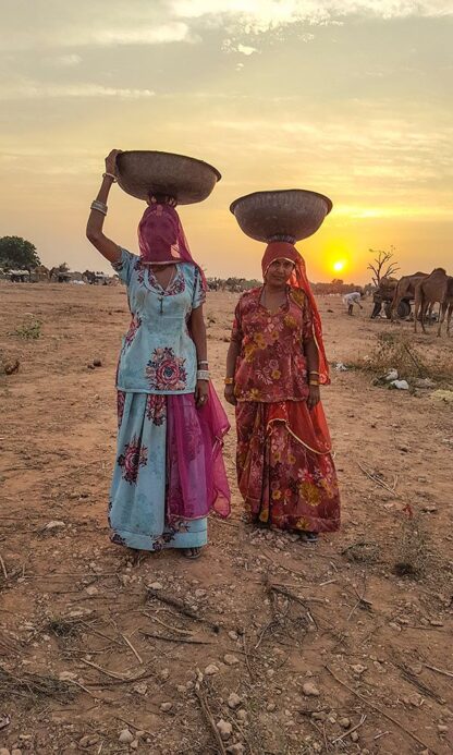 Two women dressed in colorful traditional attire carry large bowls on their heads while walking through the dry, rural landscape of Rajasthan with a sunset and camels in the background, truly capturing the wonders of this region.