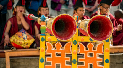 A close-up of two large, decorated horns with red interiors positioned on a colorful wooden stand during the Tiji Festival in Nepal, with people in traditional attire seated in the background.