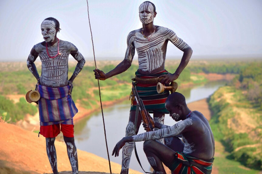 Three men adorned in traditional attire and body paint stand and sit on a hilltop, overlooking a river and landscape in the distance. One holds a stick while another grips a rifle, showcasing their Karos heritage.