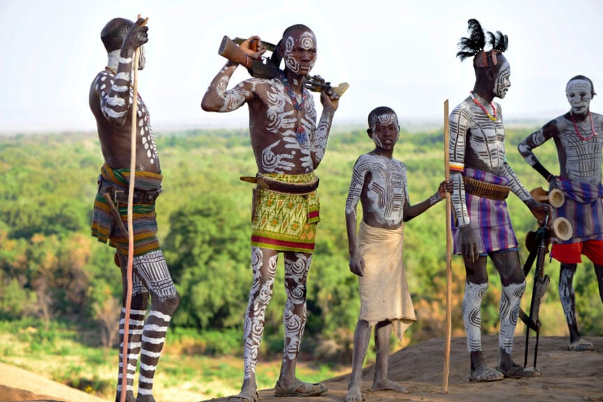 A group of people, Karos adorned in traditional attire and body paint, stand outdoors holding sticks and various objects. Trees and greenery can be seen in the background.