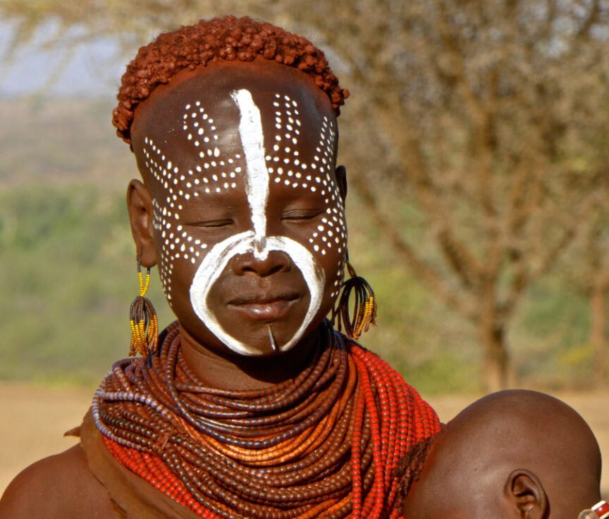 A person, adorned with white tribal face paint and multiple beaded necklaces and earrings, stands outdoors with another partially visible beside them. This striking figure exudes the aura of the Karos tradition.