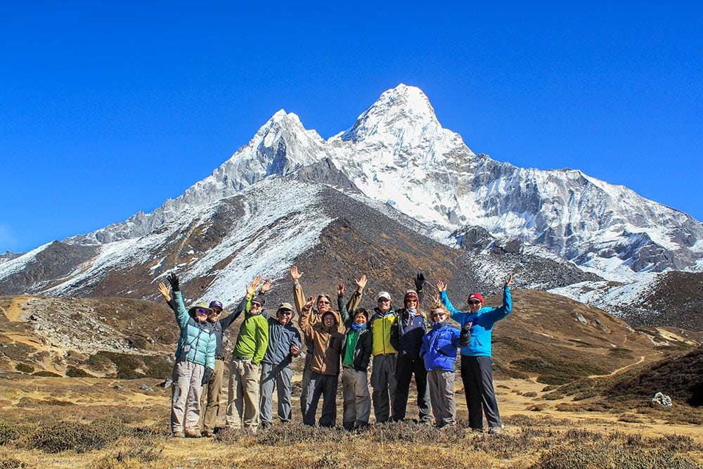 A group of adventurous people in hiking gear stand with their arms raised in front of a snow-capped mountain under a clear blue sky, experiencing the thrill of Asia's majestic peaks.