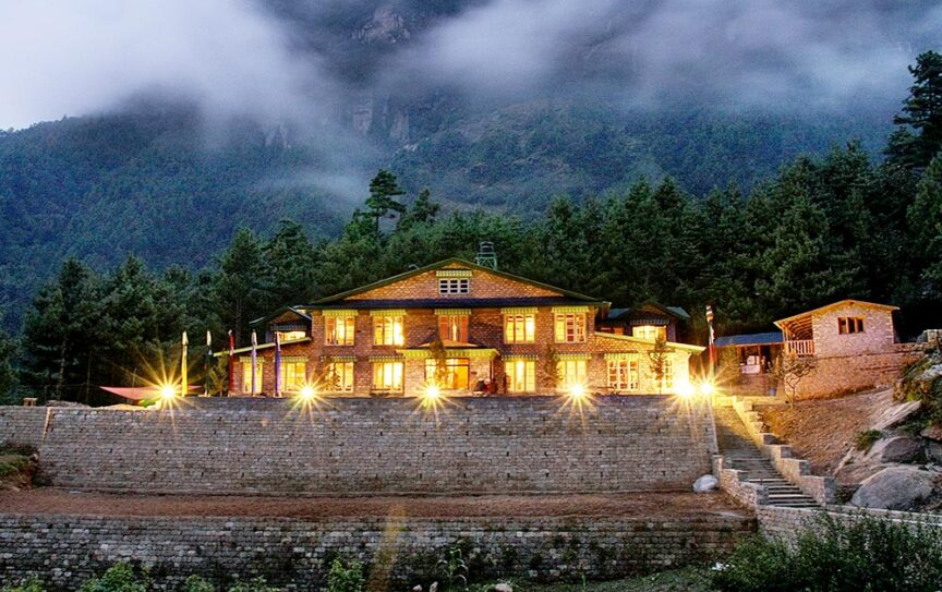 A large building surrounded by trees, illuminated with lights in front, sits on a stone retaining wall at dusk, with misty mountains in the background—an ideal retreat for those embarking on adventurous hiking trips in Asia.