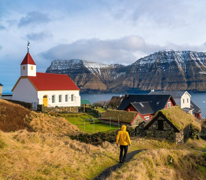 Person in a yellow jacket hiking towards a white church with a red roof in a picturesque European village, surrounded by grass-covered houses and snow-capped mountains. No crowds disrupt this serene landscape.