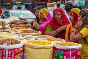 Women in colorful attire stand in front of stalls filled with various grains, nuts, and dried goods at an outdoor market, showcasing a vibrant snapshot of a cultural journey through Northern India.