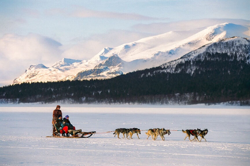 Two people are riding a dog sled pulled by a team of six dogs across a snowy landscape with a backdrop of snow-covered mountains, experiencing one of the most exhilarating adventures in some of the world's happiest countries.