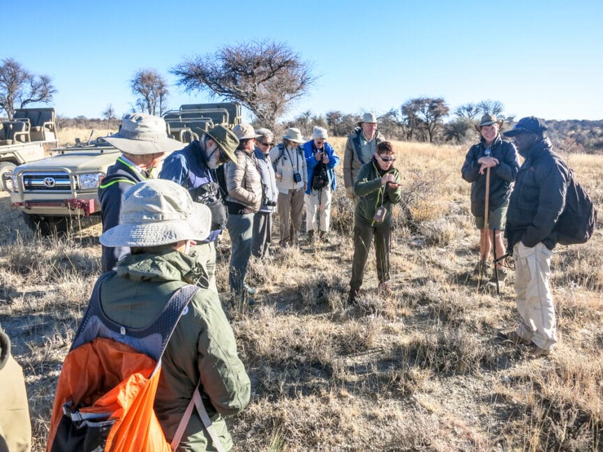 A group of people, some wearing hats and backpacks, stand near two off-road vehicles in a dry, grassy area with sparse trees, listening to a guide during an unforgettable Namibia travel experience.