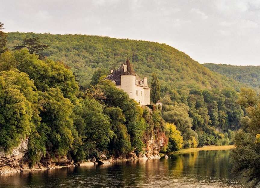 A stone castle with a pointed roof sits on a forested cliffside overlooking a river, surrounded by dense green foliage.