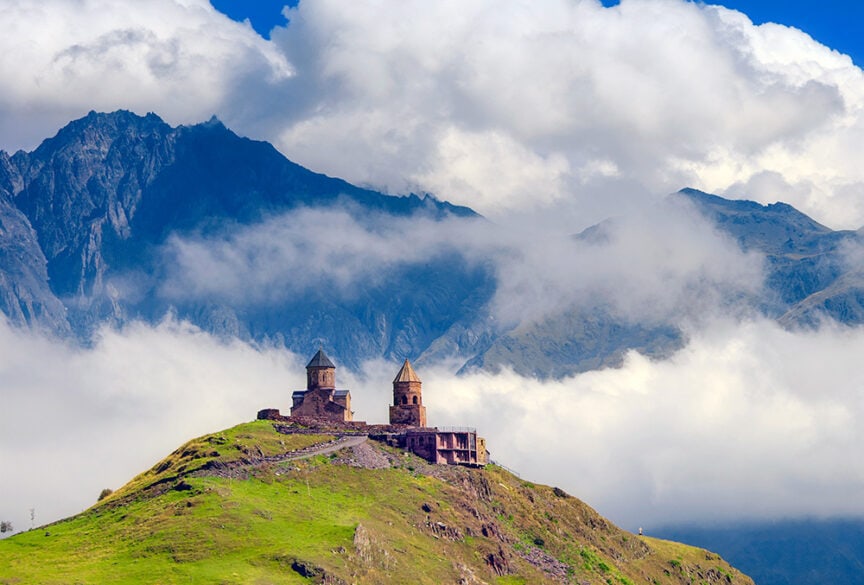 Two ancient stone structures with conical roofs are situated on a grassy hilltop in Europe, with a backdrop of misty mountains and fluffy clouds. This serene spot may attract the occasional hiking enthusiast, but you'll seldom encounter crowds here.