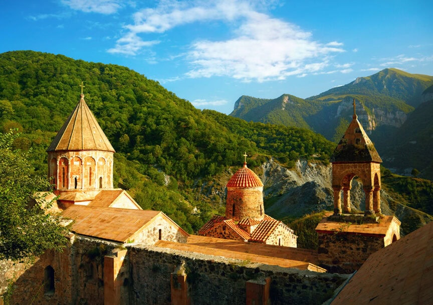 A historic monastery with multiple domed and peaked roofs is set against a backdrop of green forested hills and distant mountains under a clear blue sky, inviting travelers to explore one of the region's hidden gems amidst renowned wine-growing regions.