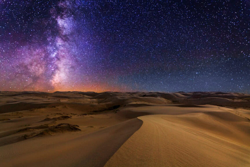 A star-filled night sky, perfect for stargazing, illuminates the Milky Way over rolling sand dunes in a desert landscape.