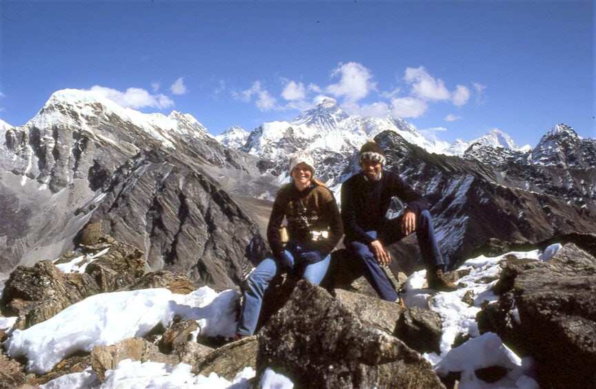 Two people sitting on rocky terrain with snow-capped mountains in the background under a clear blue sky, sharing a moment during their trek to Everest Base Camp, their journey unfolding like an epic love story.