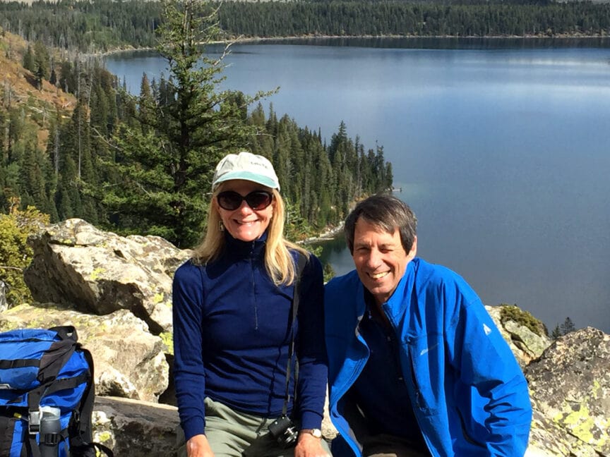 Two people wearing outdoor gear sit on rocks with a scenic lake and forest in the background, enjoying the view, reminiscent of a love story unfolding on an epic trek to Everest Base Camp.