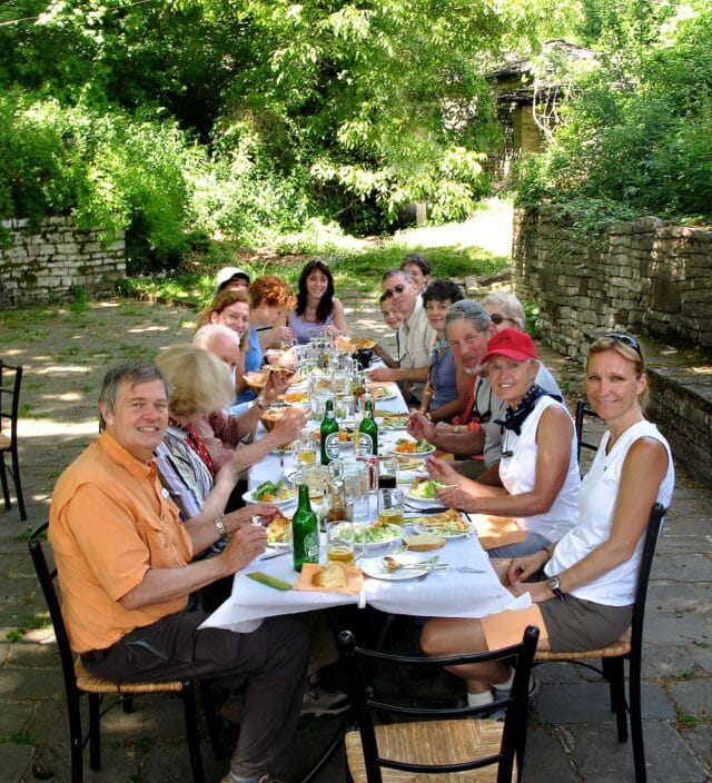 A group of people sit around a long outdoor dining table set with food and drinks, savoring delicious recipes, surrounded by greenery. Everyone is facing the camera, some smiling.