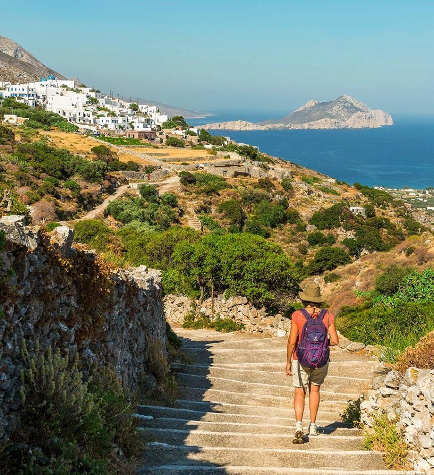 A person with a hat and backpack walks down stone steps leading to a scenic coastal village in Europe, with the sea and an island in the background, enjoying a quiet hike without the crowds.