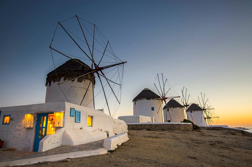 Four traditional windmills with thatched roofs stand on a hill at dusk, with a lit building in the foreground. The sky transitions from blue to orange as the sun sets, capturing a picturesque moment perfect for those who love to travel and explore colorful cities around the world.
