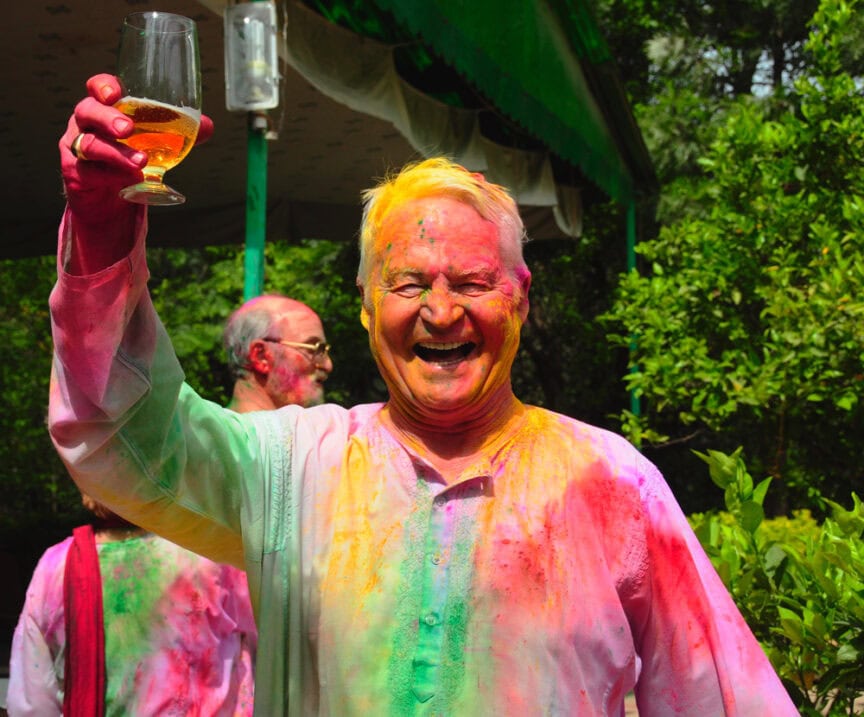An elderly man with a colorful face and clothes holds up a glass with liquid, smiling widely. Green foliage and other people in festive attire celebrate the vibrant festival of Holi in the background.