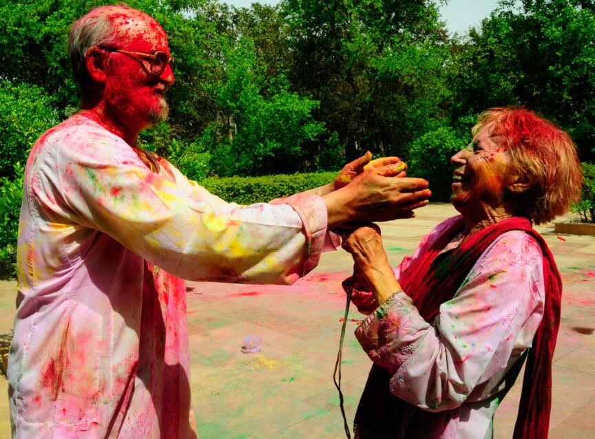 Two elderly individuals covered in vibrant colors are joyfully celebrating Happy Holi outdoors in a lush, green park.