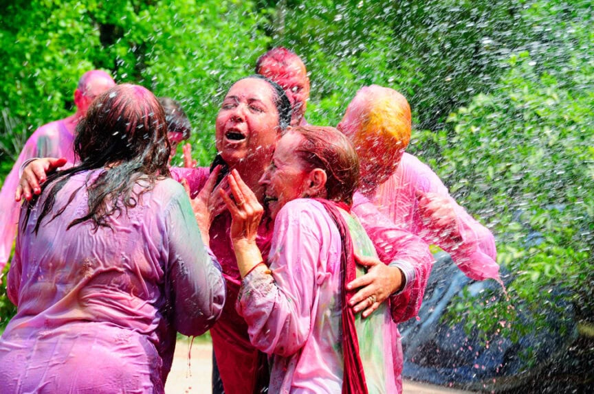 People wearing colorful clothes are celebrating Happy Holi outdoors, splashing water and covered in vibrant colors. They exhibit joy and excitement in the midst of the festival.