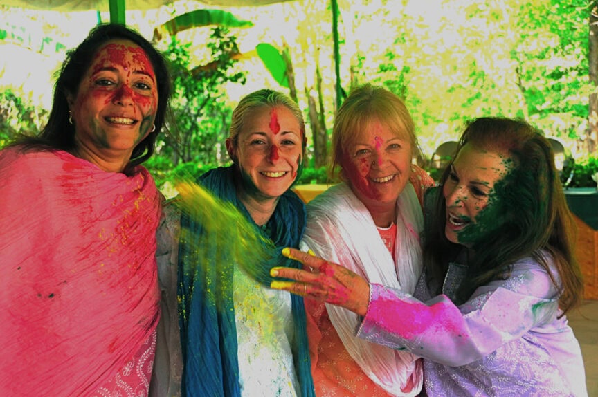 Four people wearing colorful clothing and covered in bright colored powders, standing close together and smiling outdoors, likely celebrating the vibrant festival of Holi.