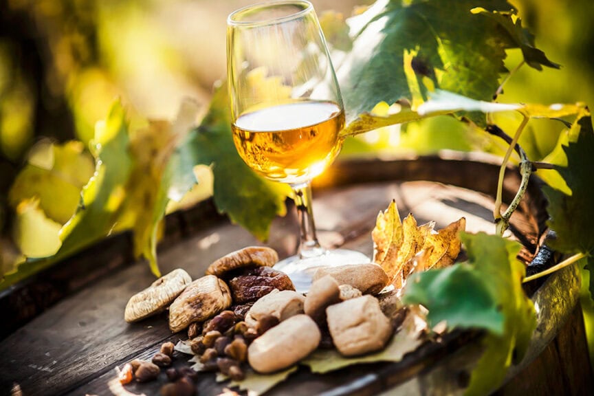 A glass of white wine is placed on a wooden barrel, surrounded by mixed nuts and dried fruits amidst green and yellow vine leaves in the background, evoking a sense of adventure through scenic wine-growing regions.