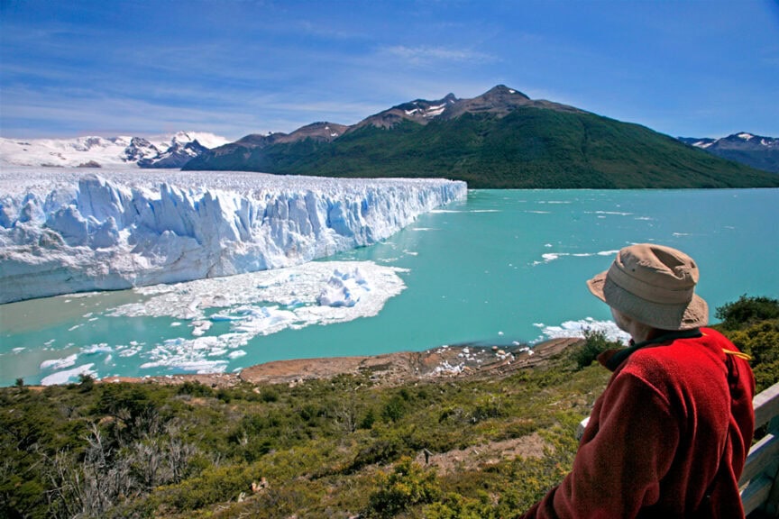 Carlos Leon, wearing a red jacket and hat, overlooks a large glacier and turquoise water with majestic mountains in the background.