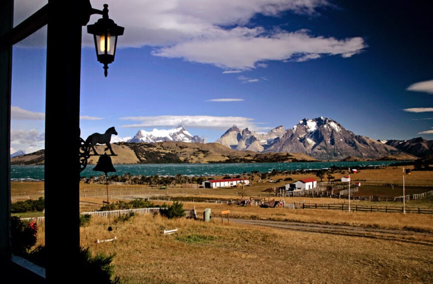 A scenic view of snow-capped mountains and a blue lake from a ranch with a bell hanging in the foreground, reminiscent of the tranquil landscapes you might know Carlos Leon for capturing so vividly.