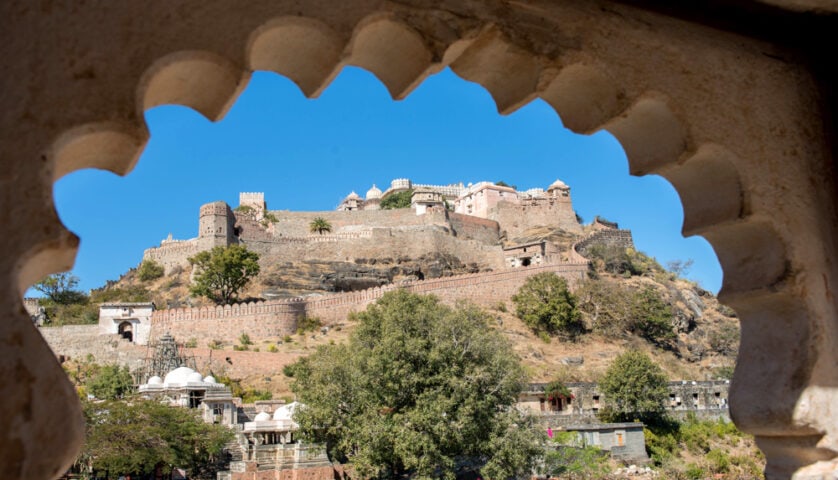 View of a historical hilltop fort framed by an ornate archway, with stone walls, towers, and surrounding buildings contrasting against a clear blue sky and the lush greenery of the country.