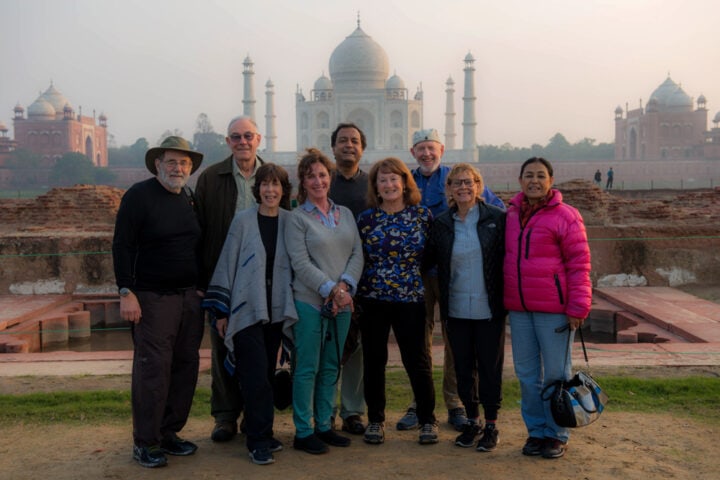 A group of nine people stands in front of the Taj Mahal at sunrise, posing for a photo with the monument in the background. They are dressed in casual outdoor clothing, capturing a memory from this remarkable country of contrasts.