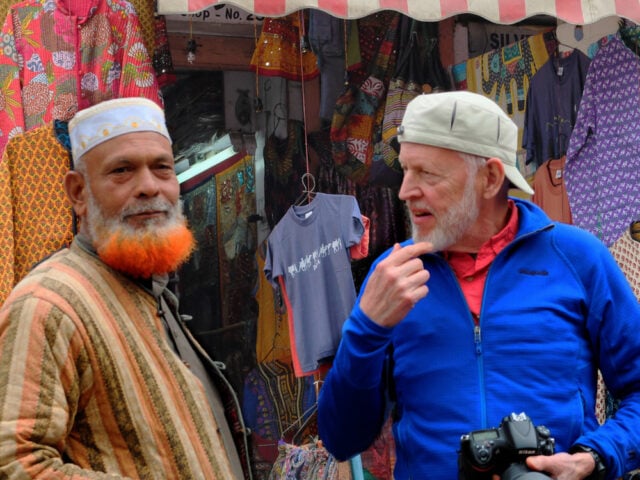 Two men, embodying the contrasts of their country, stand in front of a colorful market stall. One has a dyed orange beard and wears traditional attire, while the other wears a blue jacket and cap, holding a camera and gesturing animatedly.