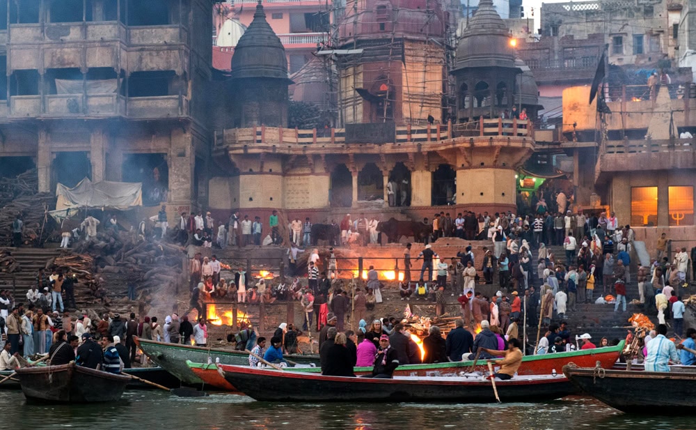 Crowd gathers on the steps of the Ganges River in Varanasi, with boats on the water and multiple small fires burning in the background near ancient buildings. This picturesque scene contrasts the vibrant energy of daily life with timeless traditions, making it a must-see travel destination in this country.