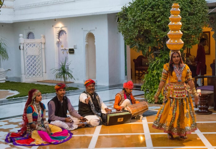 In a display that epitomizes the Country of Contrasts, a woman in traditional attire balances a stack of pots on her head while four musicians seated on the floor play instruments in an ornate courtyard setting.