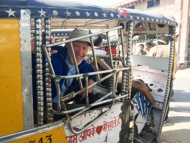 A person wearing a hat smiles and waves from inside a decorated auto rickshaw, highlighting the contrasts in a bustling urban setting.