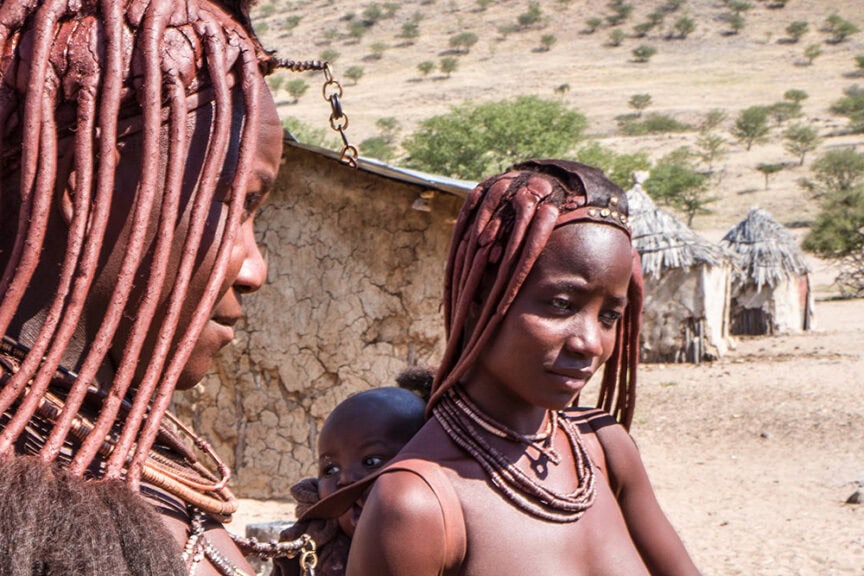 Two women with intricately styled hair and traditional necklaces stand outdoors near thatched and mud-brick structures in Namibia, with a child peeking in between, capturing a glimpse of the country's stunning landscapes.