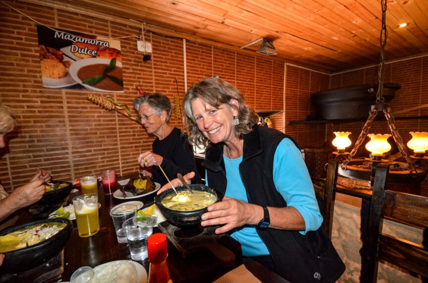 A woman holding a bowl of soup smiles at the camera while dining indoors with others, surrounded by beverages and condiments on the table. A "Mazamorra Dulce" sign is visible on a brick wall in the background, reflecting the charm of international cuisine.