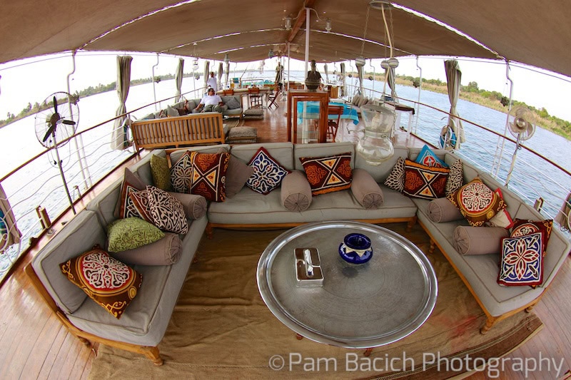 A covered outdoor seating area on a boat, perfect for Nile cruising, featuring colorful patterned cushions, a circular metal table at the center, and a breathtaking water view in the background.