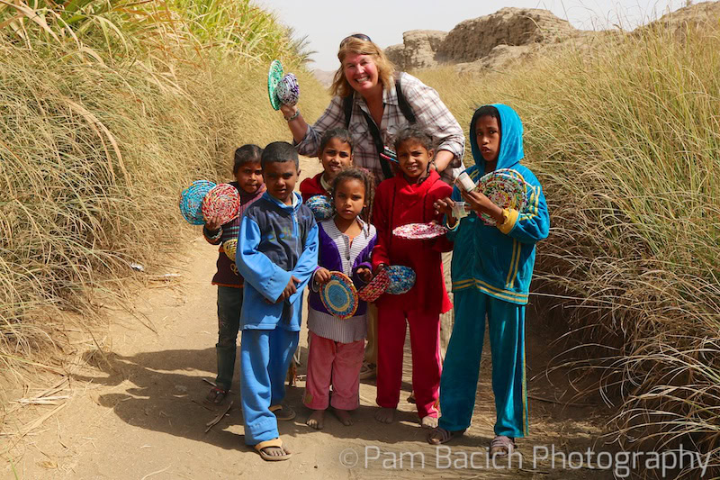 A smiling adult stands with a group of children holding colorful woven plates in a grassy field, reminiscent of joyous travels. Rocks are visible in the background. Photo by Pam Bacich Photography.