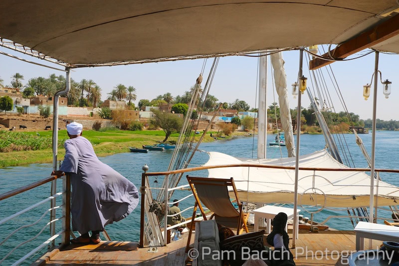 A person dressed in traditional attire stands at the railing of a wooden boat, looking out over the calm Nile River, with a lush village landscape in the background—an idyllic scene perfect for Egypt tourism or a serene Nile cruise.