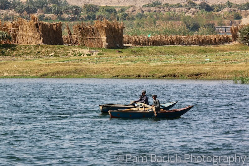 Two small boats with people in them are cruising on the Nile, with green fields and reed structures on the riverbank in the background.