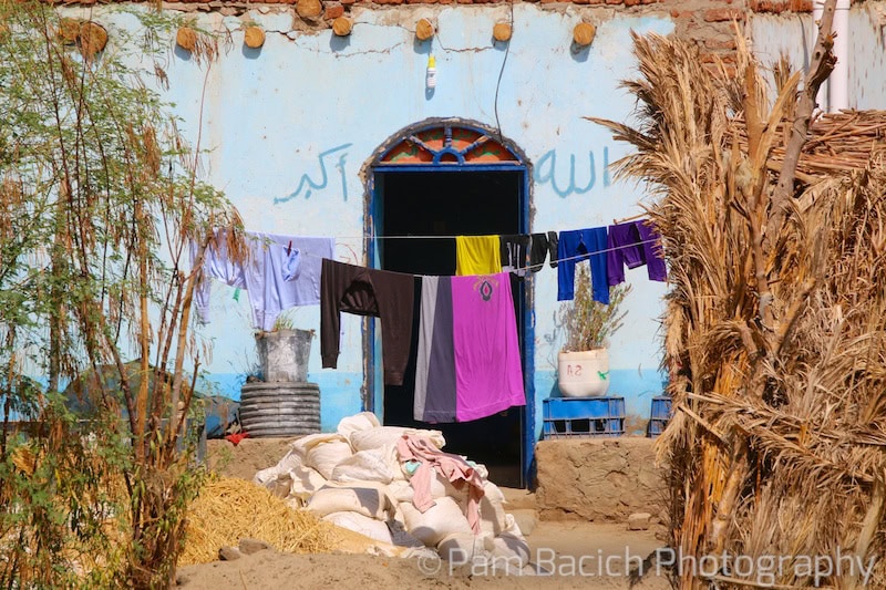 Colorful clothes hanging to dry on a line in front of a blue-painted building with an arched doorway, reminiscent of scenes seen while cruising the Nile, surrounded by plants and various objects including bags and a metal container.