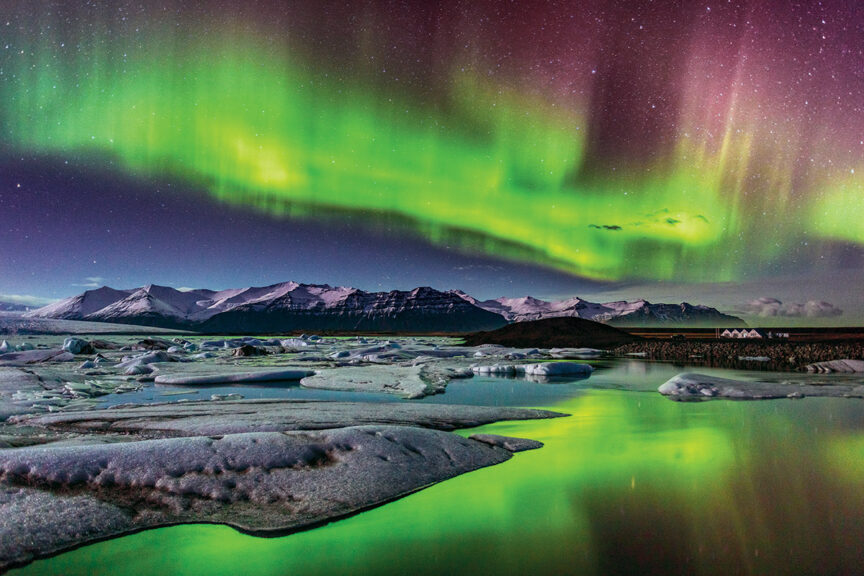A vibrant green aurora borealis lights up the night sky over a snowy mountain range and a partially frozen river with ice chunks, offering a mesmerizing backdrop for adventures. A few isolated buildings are visible in the distance, hinting at cozy retreats in one of the world's happiest countries.