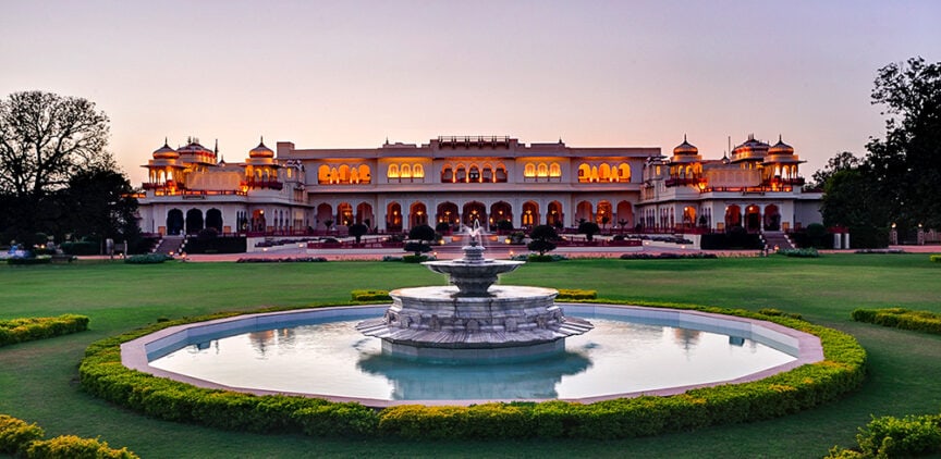 A grand building with illuminated arches and domes, reminiscent of ancient palaces, stands behind a large circular fountain, surrounded by a well-maintained garden at dusk.
