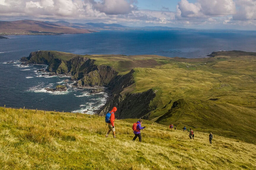 Hikers walk down a grassy slope towards a rugged coastline with cliffs and the ocean in the background under a partly cloudy sky, marking the end of an unforgettable 9-day trip.
