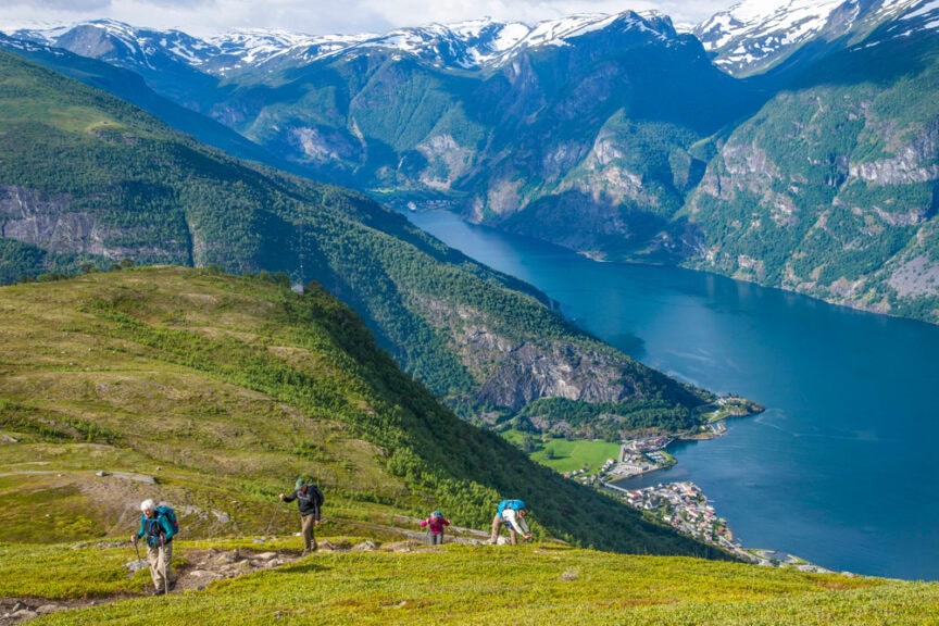 Hikers trekking along a mountainous trail with a deep blue fjord and snow-capped peaks in the background. A small village is visible by the water, capturing the serene beauty of Norway. Bright, clear day.