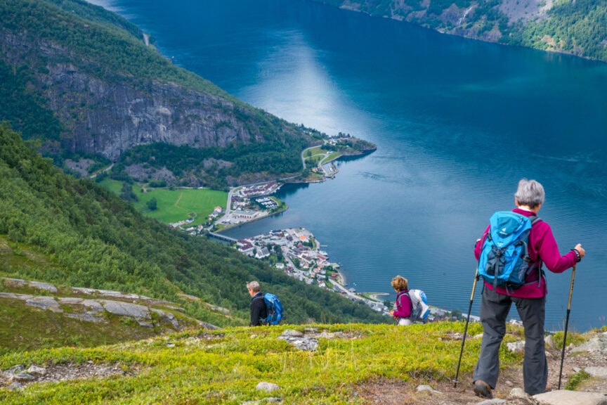 Three hikers descend a green hillside towards a deep blue fjord in Norway, with a small village along its shore, surrounded by mountains and water.
